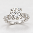 A classic solitaire engagement ring featuring a round brilliant-cut diamond center stone in a four-prong setting, with a split shank adorned with small pavé diamonds, shown on a light gray background.
